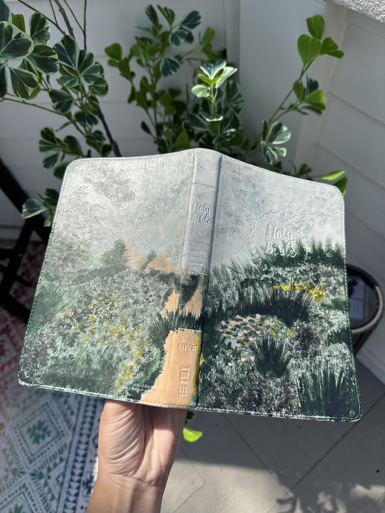 May include: An open Bible with a painted landscape design. The cover features a detailed illustration of a path winding through a field of green and yellow flowers. The spine is embossed with the words "Holy Bible".