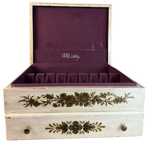 May include: Open, vintage jewelry box with a distressed white finish. The interior is lined with a deep purple fabric and features compartments for storage. The exterior is decorated with floral designs in shades of brown and gold. The box has a drawer with a round knob.