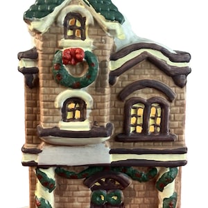 May include: A ceramic Christmas house figurine with a green roof and brick facade. The house features a wreath, garland, and snow accents. Windows are illuminated, and the base is painted blue.
