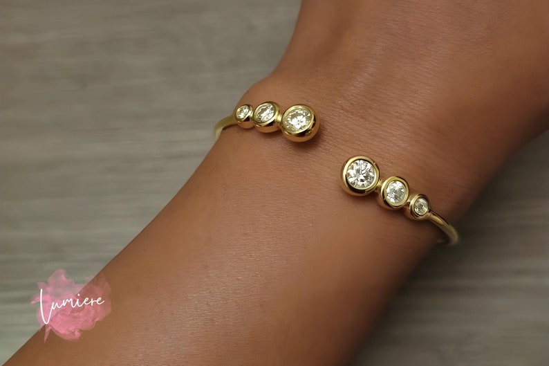 May include: A gold bangle bracelet with five clear round stones set in the metal. The bracelet is open and has a curved shape.