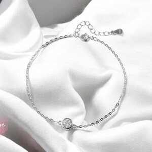 May include: A delicate silver anklet with a small round clear crystal charm. The anklet is on a white fabric background.