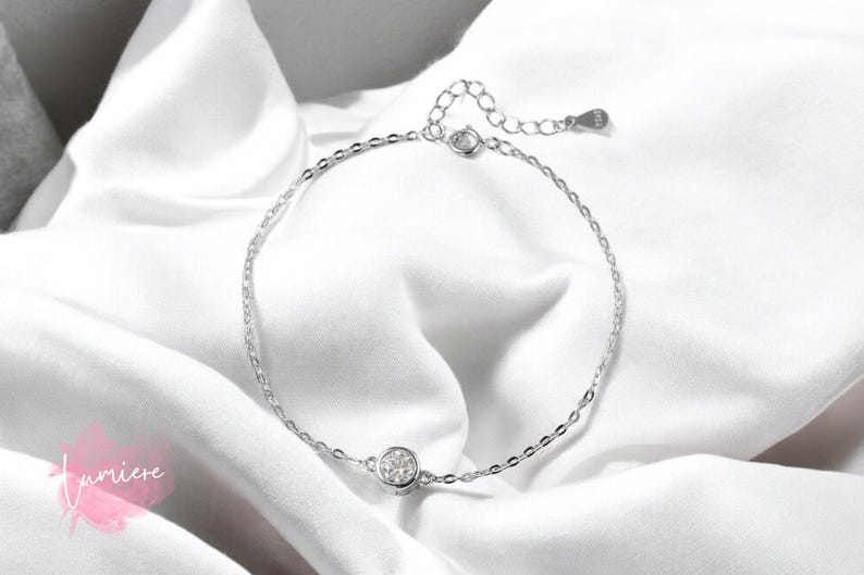 May include: A delicate silver anklet with a small round clear crystal charm. The anklet is on a white fabric background.