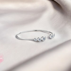 May include: A silver bangle bracelet with five clear round stones set in the metal. The bracelet is open on one side and has a curved shape.