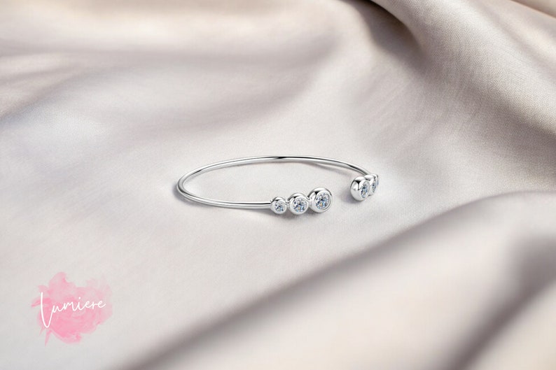May include: A silver bangle bracelet with five clear round stones set in the metal. The bracelet is open on one side and has a curved shape.