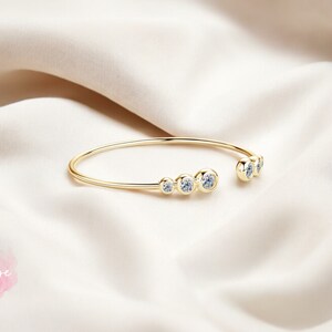 May include: A gold bangle bracelet with a row of clear round stones. The bracelet is open on one side and has a simple design.