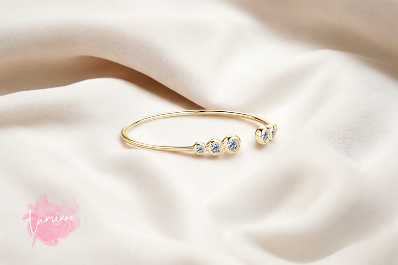 May include: A gold bangle bracelet with a row of clear round stones. The bracelet is open on one side and has a simple design.