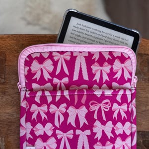 May include: A pink and white bow-patterned tablet sleeve with a zipper closure. The sleeve has a pocket on the front and a pink zipper. The text on the label reads "It's Tex Jess". An e-reader is partially visible inside the sleeve.