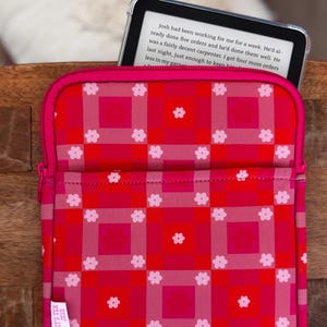 May include: A red and pink patterned e-reader sleeve with a zipper closure. The sleeve features a grid design with white flower accents and the text "IT'S TEX JESS". An e-reader is partially visible inside the sleeve.