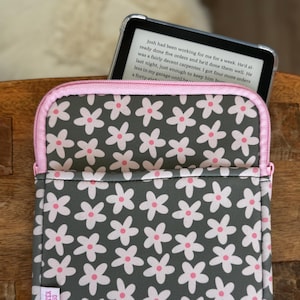 May include: Gray and pink floral pattern e-reader sleeve with a pink zipper and trim. The sleeve has a repeating pattern of white flowers with pink centers on a gray background. The text "IT'S TEX JESS" is visible on the lower left corner.