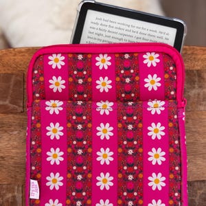 May include: A pink Kindle case with a daisy and floral pattern. The case has a zipper closure and a small white tag that says "IT'S TEX JESS". A Kindle is partially visible inside the case.