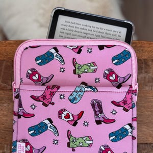 May include: Pink e-reader case with a zipper closure, featuring a repeating pattern of colorful cowboy boots and stars. The case holds an e-reader displaying text. The brand name "IT'S TEX JESS" is visible on the side.