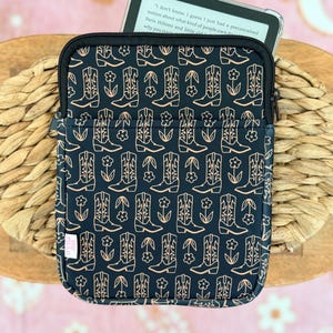 May include: Black and tan ebook reader case with a zipper closure. The case features a repeating pattern of cowboy boots and flowers. A digital reader is partially visible inside the case, displaying text.