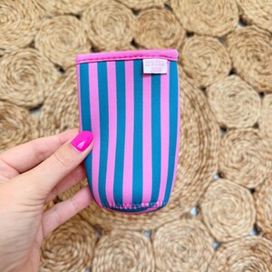 May include: A colorful drink sleeve with vertical stripes in teal and pink. The top and bottom edges are trimmed in pink. A small white tag with text is visible on the side. The sleeve is held against a textured, woven background.