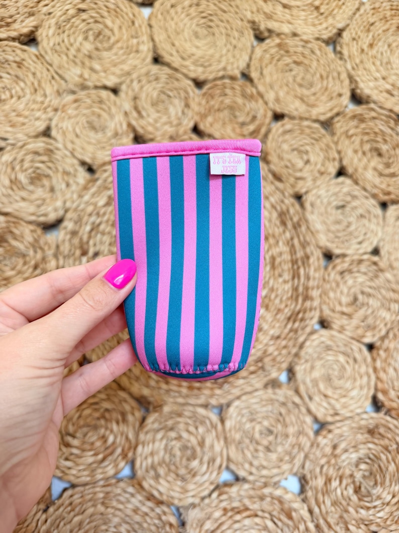 May include: A colorful drink sleeve with vertical stripes in teal and pink. The top and bottom edges are trimmed in pink. A small white tag with text is visible on the side. The sleeve is held against a textured, woven background.