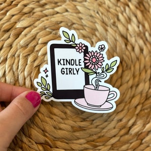 May include: A white sticker with a black outline of a Kindle e-reader, a pink teacup with steam, and a pink flower with green leaves. The text "Kindle Girly" is on the e-reader.