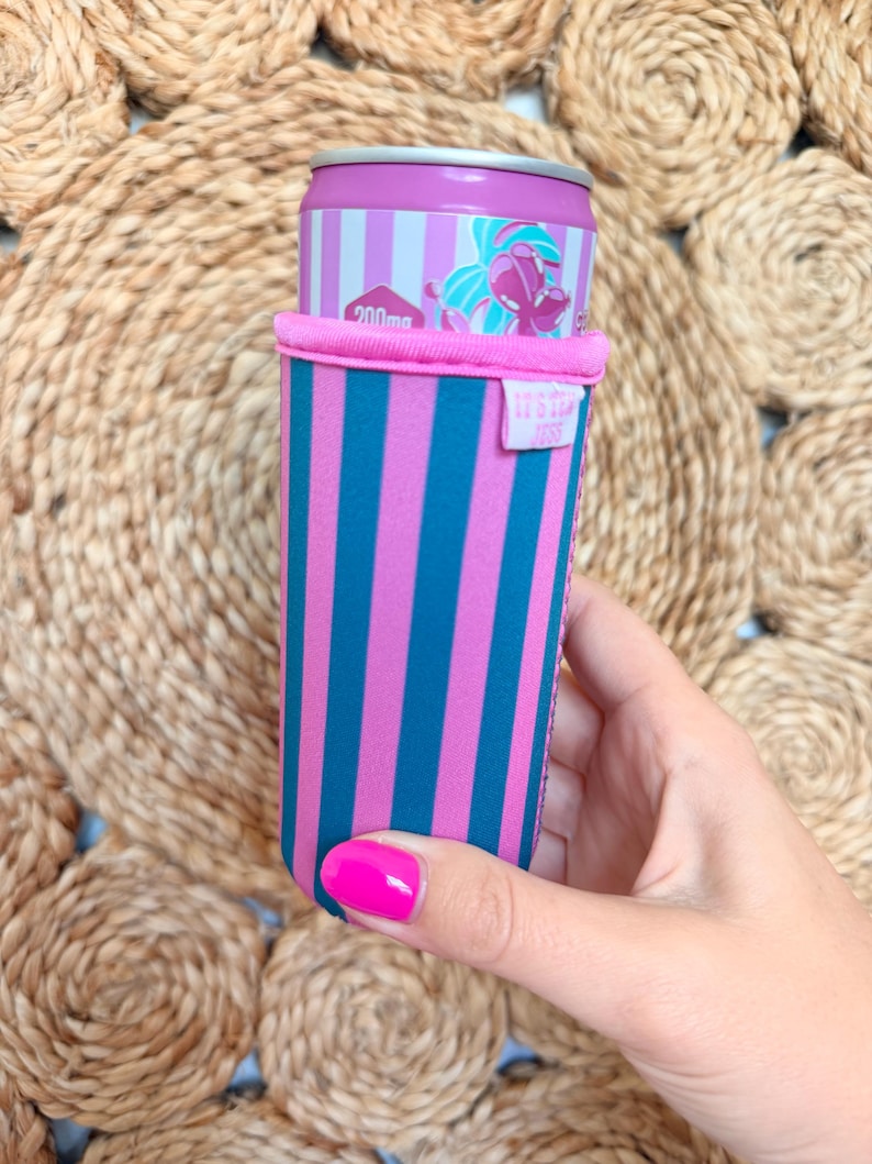 May include: A pink and teal striped can cooler holding a can with a pink and white striped design. The can cooler has a pink top and a small tag with white text. The background is a woven, circular mat.