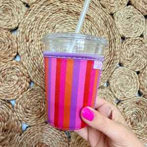 May include: A reusable drink sleeve with vertical stripes in shades of pink, orange, and purple. The sleeve is wrapped around a clear plastic cup with a straw. The text "It's just" is visible on the sleeve.