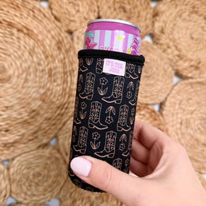 May include: A black can cooler featuring a repeating pattern of cowboy boots and flowers in a copper color. The text "IT'S TEX JESS" is printed on a white label. A pink and purple can is inside the cooler.