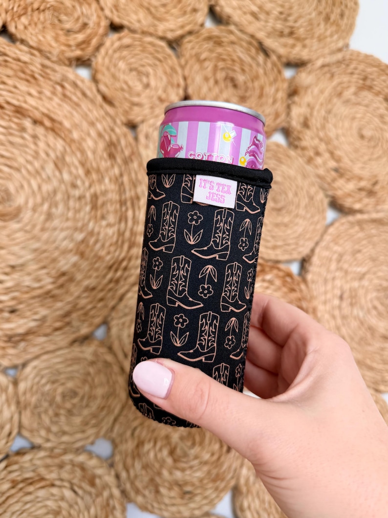 May include: A black can cooler featuring a repeating pattern of cowboy boots and flowers in a copper color. The text "IT'S TEX JESS" is printed on a white label. A pink and purple can is inside the cooler.