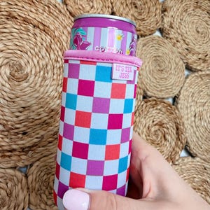 May include: A colorful drink can cooler with a checkered pattern in shades of pink, blue, and red. The cooler has a pink rim and a small tag that reads "IT'S TEX JESS". A can of Cotton soda is inside the cooler.