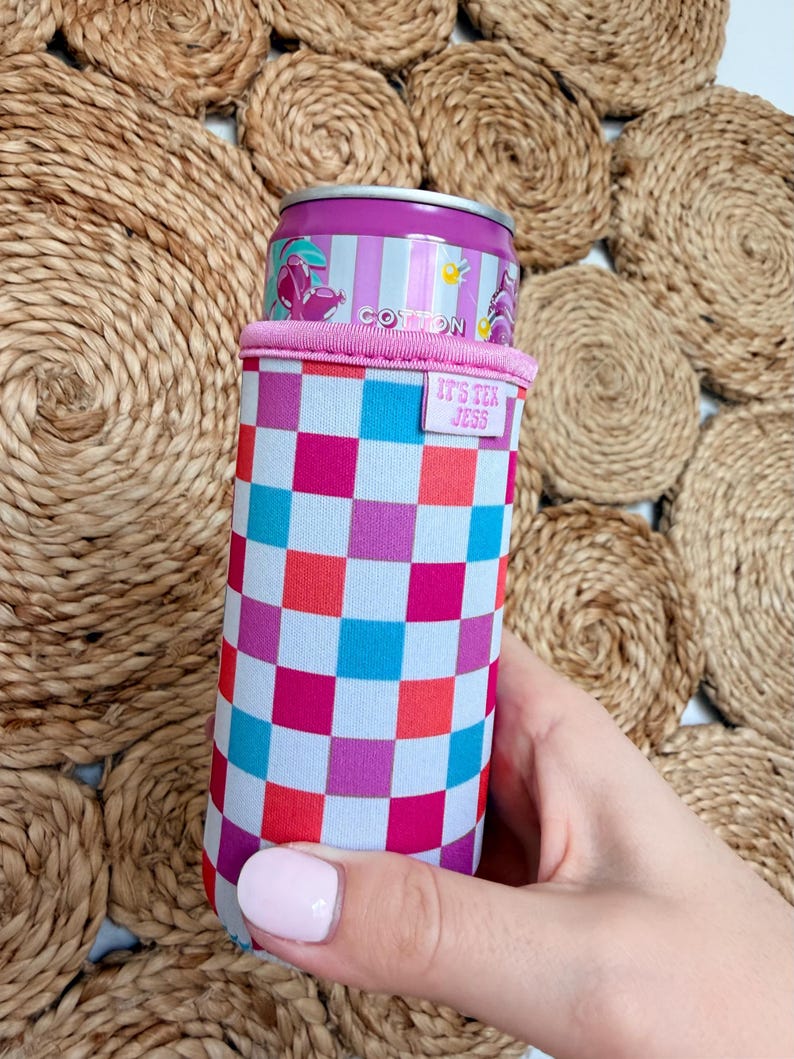 May include: A colorful drink can cooler with a checkered pattern in shades of pink, blue, and red. The cooler has a pink rim and a small tag that reads "IT'S TEX JESS". A can of Cotton soda is inside the cooler.