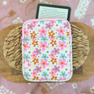 May include: A rectangular e-reader sleeve featuring a repeating floral pattern in shades of pink, orange, light blue, and white on a beige background. The sleeve has a light pink zipper closure and a small white fabric tag on the lower left side.