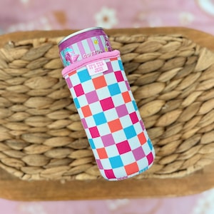 May include: A colorful can cooler with a checkerboard pattern in pink, blue, orange, and white. The cooler has a pink rim and the text "IT'S TEA JESS". A can of tea is inside the cooler.