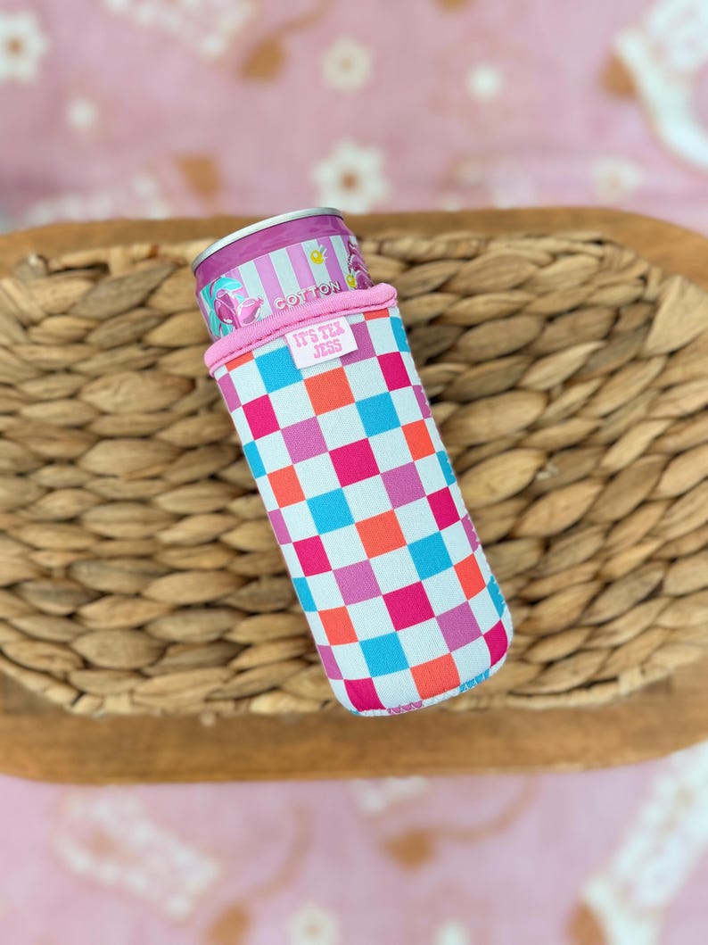 May include: A colorful can cooler with a checkerboard pattern in pink, blue, orange, and white. The cooler has a pink rim and the text "IT'S TEA JESS". A can of tea is inside the cooler.