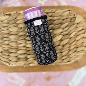 May include: A black can cooler with a pattern of cowboy boots and flowers in a copper color. The cooler holds a pink and silver can. The text "IT'S TEX JESS" is on a small white tag.