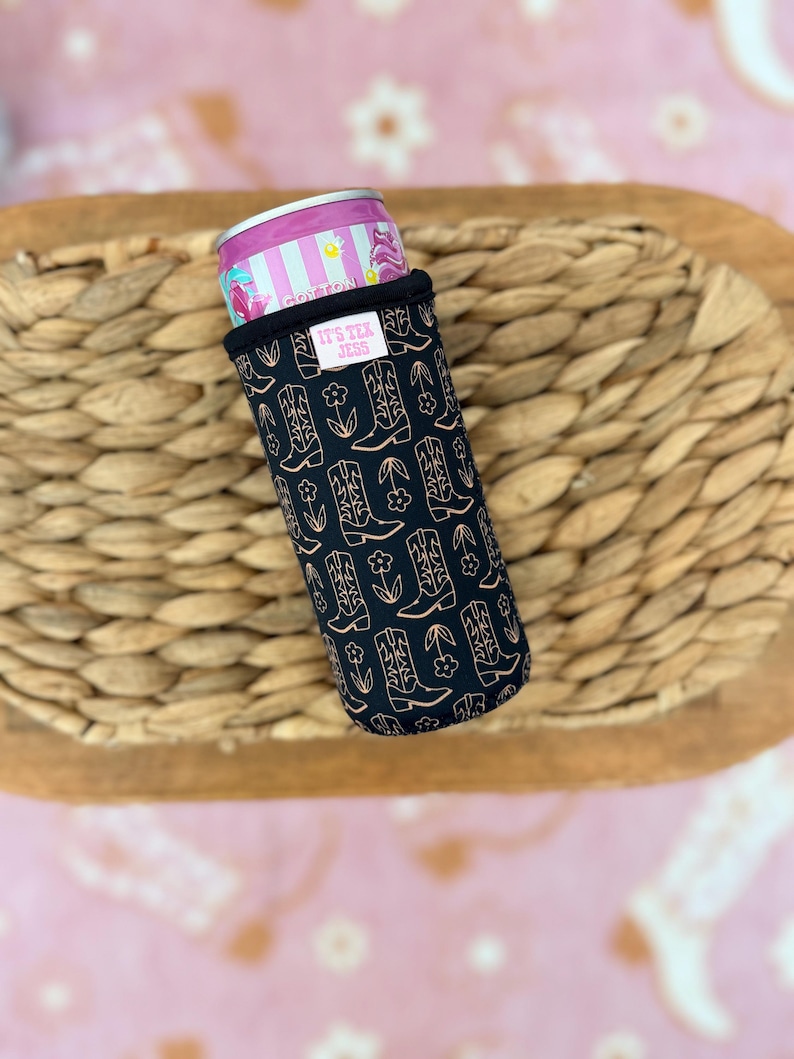 May include: A black can cooler with a pattern of cowboy boots and flowers in a copper color. The cooler holds a pink and silver can. The text "IT'S TEX JESS" is on a small white tag.