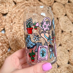 May include: A clear glass can-shaped drinking glass decorated with gardening-themed illustrations. The design includes a pink watering can, flower pots, boots, and a packet of flower seeds. The glass is held against a woven background.