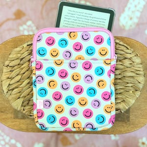 May include: A rectangular tablet sleeve featuring a repeating pattern of colorful smiley faces in pink, orange, purple, and blue on a white background. The sleeve has a light pink zipper closure and contains a tablet displaying visible text.