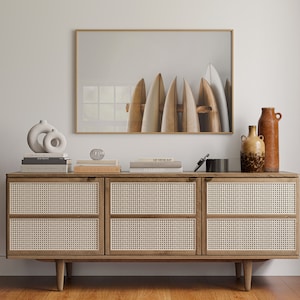 May include: A light wood cabinet with four rattan doors and a gold metal handle on each door. The cabinet is sitting on a wooden floor. Above the cabinet is a framed print of surfboards against a white background. The print has a gold frame. There are books and a small glass orb on top of the cabinet. Two ceramic vases are on the right side of the cabinet.