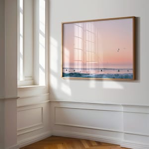 May include: A framed print depicting a serene ocean scene at sunset. Surfers are seen riding waves, with birds flying overhead. The artwork is encased in a gold-colored frame and hangs on a white wall.