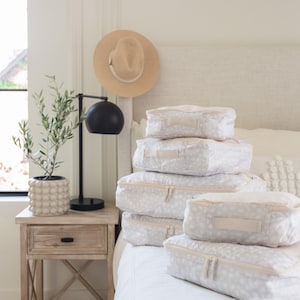 May include: A stack of six white and beige patterned travel storage cubes with zippers on a white bed. The cubes are stacked on top of each other and are arranged in a pyramid shape.
