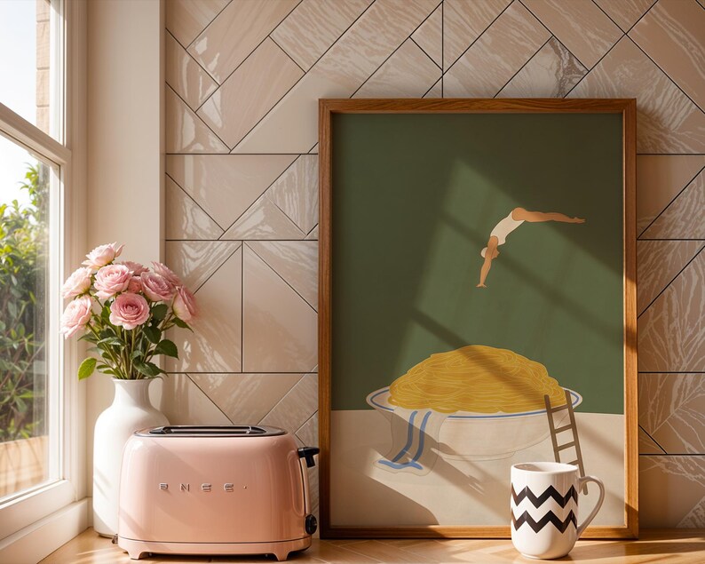 May include: A pink retro-style toaster sits next to a vase of pink roses and a framed art print. The art print features a person diving into a bowl of spaghetti. A black and white zig-zag patterned mug is also visible.