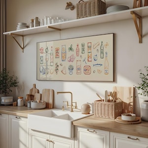 May include: A framed print featuring colorful illustrations of various food items, including pasta, wine bottles, and fish. The artwork is displayed above a white kitchen sink with a gold faucet, wooden countertops, and white cabinets. A wicker basket holds utensils.