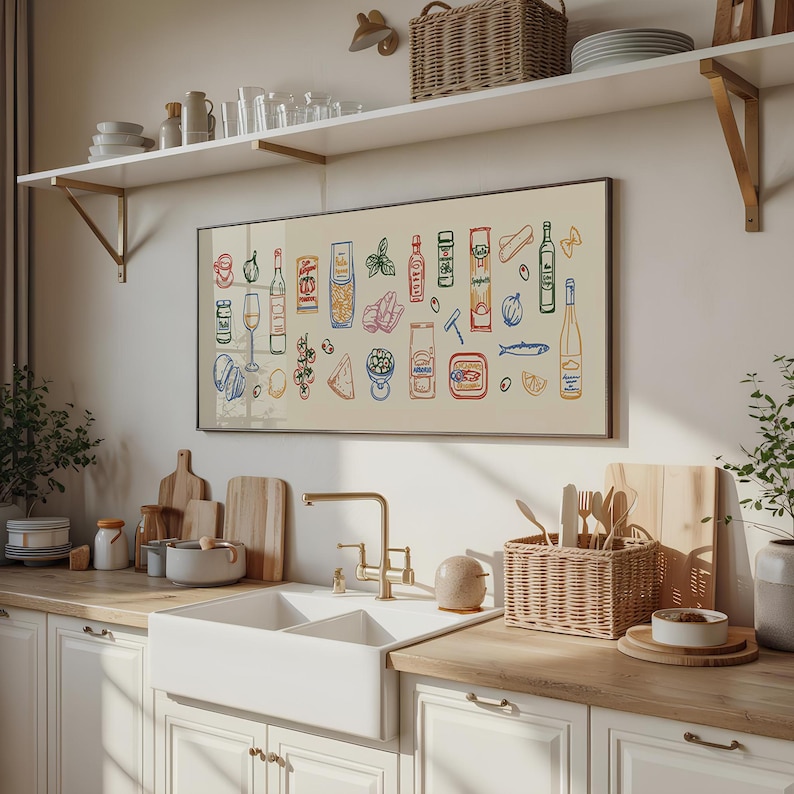 May include: A framed print featuring colorful illustrations of various food items, including pasta, wine bottles, and fish. The artwork is displayed above a white kitchen sink with a gold faucet, wooden countertops, and white cabinets. A wicker basket holds utensils.