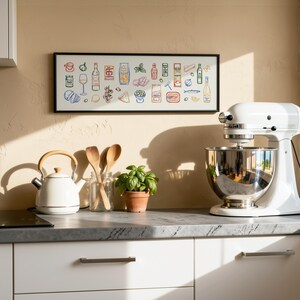 May include: A white stand mixer with a stainless steel bowl sits on a countertop next to a white kettle and a framed print of food illustrations. Wooden spoons and a small potted plant are also on the counter. The kitchen has white cabinets.