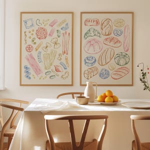 Puede incluir: Dos impresiones enmarcadas, una con una ilustraci&oacute;n colorida de varias formas de pasta y la otra con una ilustraci&oacute;n colorida de diferentes tipos de pan. Las impresiones est&aacute;n colgadas en una pared blanca sobre una mesa de comedor puesta con un mantel blanco, dos sillas de madera, un taz&oacute;n y un plato de naranjas.