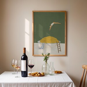May include: A framed art print depicts a person diving into a bowl of yellow food. A bottle of red wine, wine glasses, and a wooden tray with food are on a white tablecloth. A clear vase with flowers and a clear carafe are also on the table.