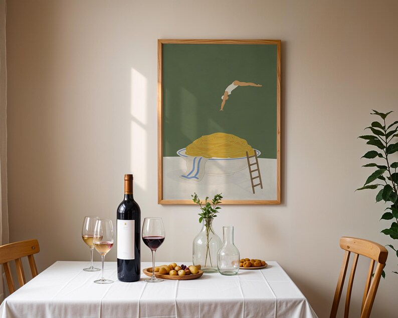 May include: A framed art print depicts a person diving into a bowl of yellow food. A bottle of red wine, wine glasses, and a wooden tray with food are on a white tablecloth. A clear vase with flowers and a clear carafe are also on the table.