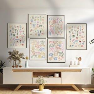 Puede incluir: Cinco ilustraciones coloridas de alimentos y bebidas enmarcadas y colgadas en una pared blanca sobre un estante de madera claro. Las ilustraciones incluyen una variedad de quesos, panes, vino y c&oacute;cteles.