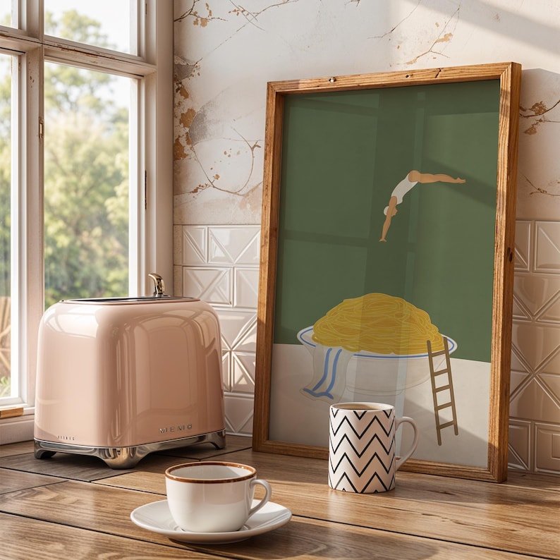 May include: A pink toaster with chrome accents sits on a wooden countertop next to a framed art print. The art features a person diving into a bowl of spaghetti. A white mug with a black zigzag pattern and a teacup are also on the counter.