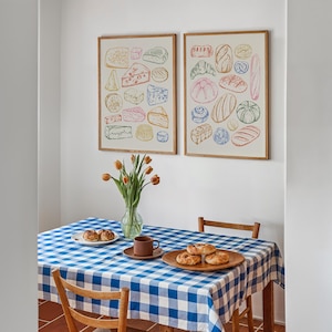 Puede incluir: Dos ilustraciones enmarcadas de diferentes tipos de alimentos. La ilustraci&oacute;n de la izquierda muestra una variedad de quesos, mientras que la ilustraci&oacute;n de la derecha muestra una variedad de panes. Las ilustraciones son de estilo vintage y est&aacute;n impresas en colores pastel. Las ilustraciones est&aacute;n colgadas en una pared blanca sobre un mantel a cuadros azul y blanco.