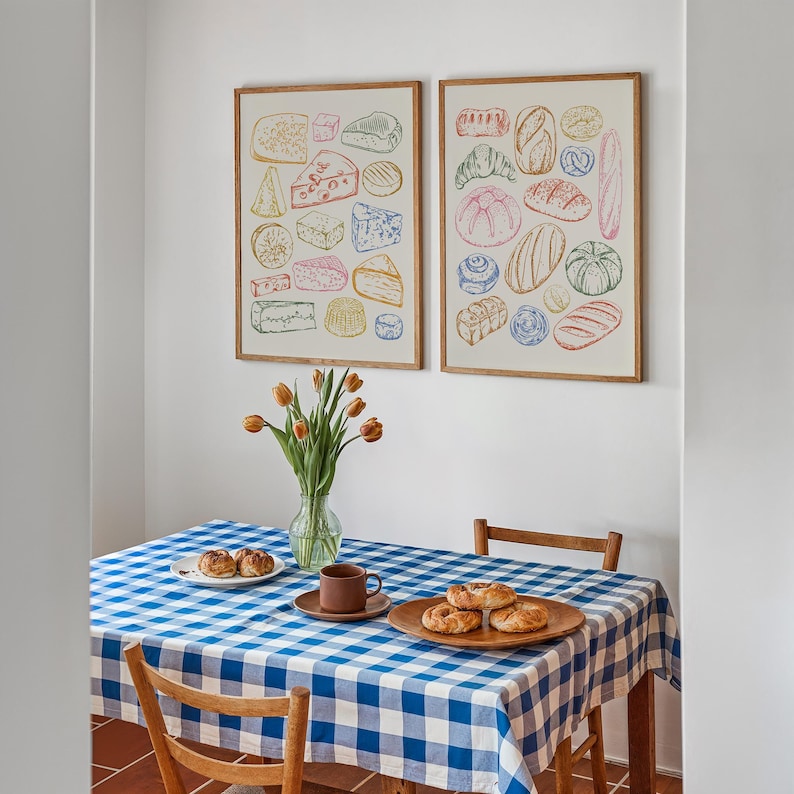 Puede incluir: Dos ilustraciones enmarcadas de diferentes tipos de alimentos. La ilustraci&oacute;n de la izquierda muestra una variedad de quesos, mientras que la ilustraci&oacute;n de la derecha muestra una variedad de panes. Las ilustraciones son de estilo vintage y est&aacute;n impresas en colores pastel. Las ilustraciones est&aacute;n colgadas en una pared blanca sobre un mantel a cuadros azul y blanco.