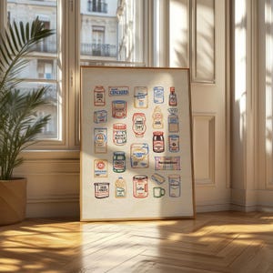 May include: A framed print featuring a colorful illustration of various pantry staples. The artwork includes images of pasta, crackers, peanut butter, and coffee, among other items. The print is displayed in a room with a window and a potted plant.