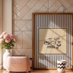 Puede incluir: Una tostadora SMEG rosa se encuentra junto a una ventana con un jarr&oacute;n de rosas rosas. Una impresi&oacute;n enmarcada presenta un fondo beige con una ilustraci&oacute;n en blanco y negro de una comida. Una taza blanca con zigzags negros est&aacute; frente a la impresi&oacute;n.