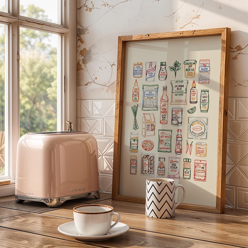 May include: A pink toaster with chrome accents sits on a wooden surface next to a framed print of food items. A white mug with a black zigzag pattern and a teacup and saucer are also on the table. The print is in a wooden frame.