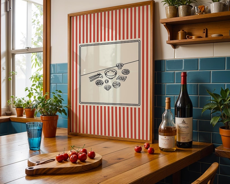 Puede incluir: Una obra de arte enmarcada con un fondo a rayas rojas y blancas y una ilustraci&oacute;n en blanco y negro de alimentos. Una tabla de cortar de madera con cerezas, un vaso azul y botellas de vino est&aacute;n sobre una mesa de madera.